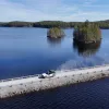 Ein See, über den eine Schotterstraße führt. Auf der Straße fährt ein weißes Auto. Im See stehen Bäume