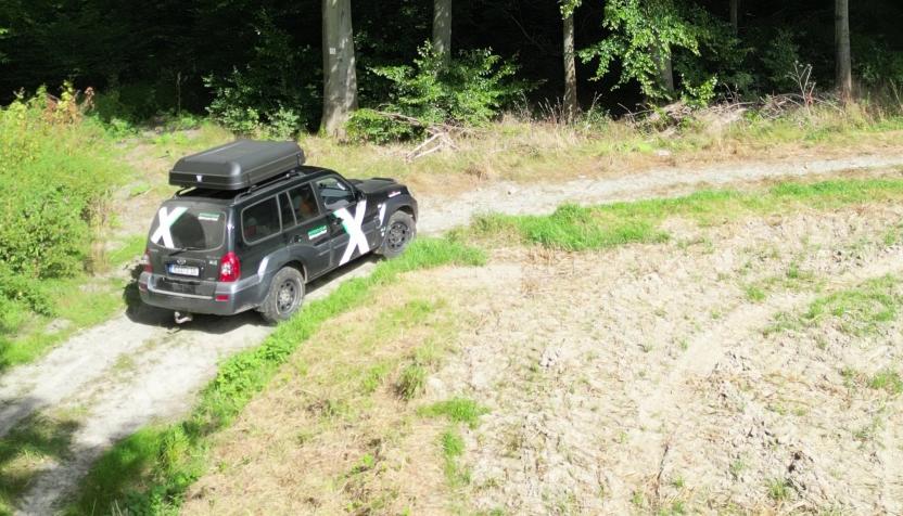 Schwarzer Geländewagen mit Dachbox und weißem X  auf Beifahrertür fährt einen Feldweg entlang. Links befindet sich ein Wald und rechts eine Wiese 