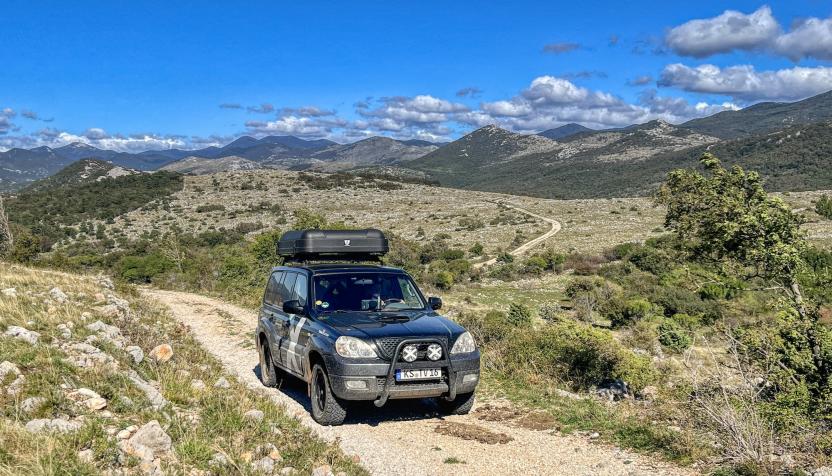 Ein schwarzer Geländewagen mit Dachbox fährt auf einem steinigen Pfad durch eine hügelige, trockene Landschaft. Im Hintergrund erstrecken sich Berge unter einem blauen Himmel mit einigen Wolken.