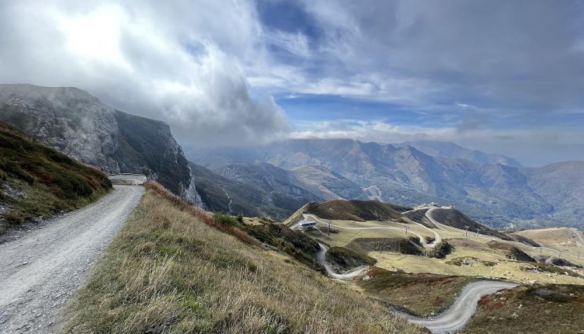 Ein kurviger Schotterweg auf der LGKS führt entlang eines steilen Berghangs, mit weiten Tälern, gewundenen Straßen und Wolken, die über die Berggipfel ziehen.