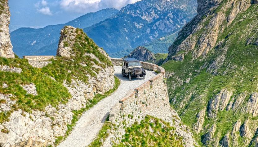 Ein Geländewagen fährt auf einem schmalen, kurvigen Bergweg entlang einer steilen Klippe, umgeben von grünen Felsen und majestätischen Bergketten.