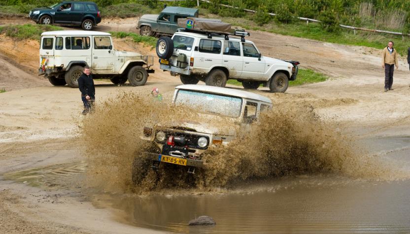 Ein weißer Geländewagen mit gelben Nummernschild fährt durch eine schlammige Wasserpfütze, spritzt dabei Wasser und Matsch auf. Im Hintergrund fahren weitere Geländewagen.