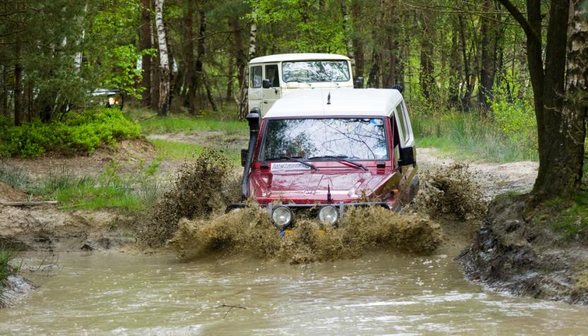 Ein roter Geländewagen mit weißem Dach fährt durch eine schlammige Pfütze im Wald und spritzt Wasser auf. Ein weiteres weißes Fahrzeug folgt dahinter.