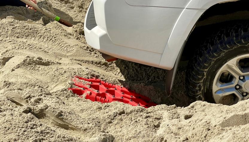 Ein Autoreifen ist im tiefen Sand eingesunken, während eine rote Traktionshilfe darunter platziert wird. Eine Person gräbt mit einer Schaufel.