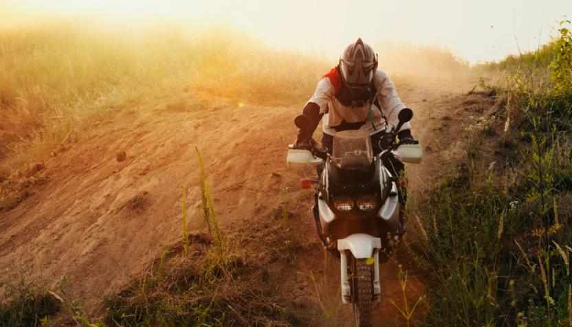 Ein Motorradfahrer fährt vorsichtig einen sandigen Hang hinauf, umgeben von trockenem Gras, in warmem, goldenem Licht der untergehenden Sonne.
