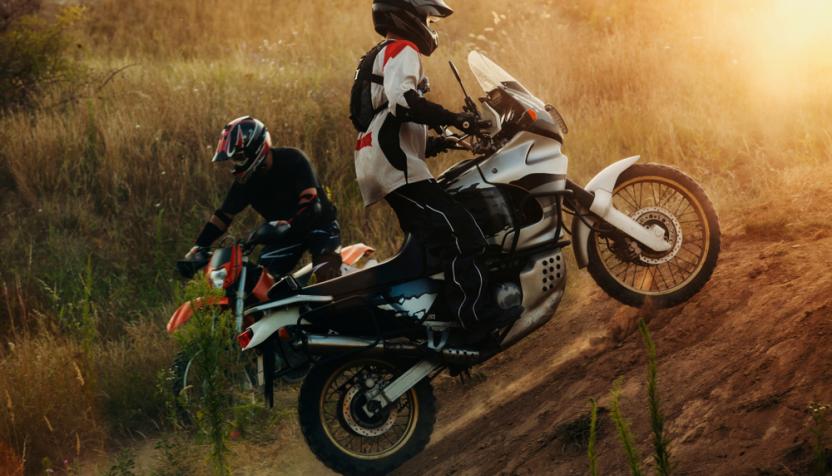 Zwei Motorradfahrer fahren eine steile, staubige Böschung hinauf in einer trockenen, grasbewachsenen Landschaft bei Sonnenuntergang.