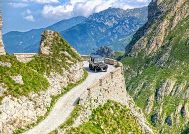 Ein Geländewagen fährt auf einem schmalen, kurvigen Bergweg entlang einer steilen Klippe, umgeben von grünen Felsen und majestätischen Bergketten.