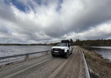Weißes Offroadauto mit Dachzelt fährt auf einer Schotterstraße über einen See