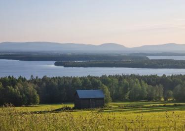 Picturesque landscape featuring a barn, lush forest, and a serene lake under a clear sky.