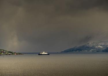 A ferry traversing the serene waters of Harstad, surrounded by dramatic skies and coastline in Norway.
